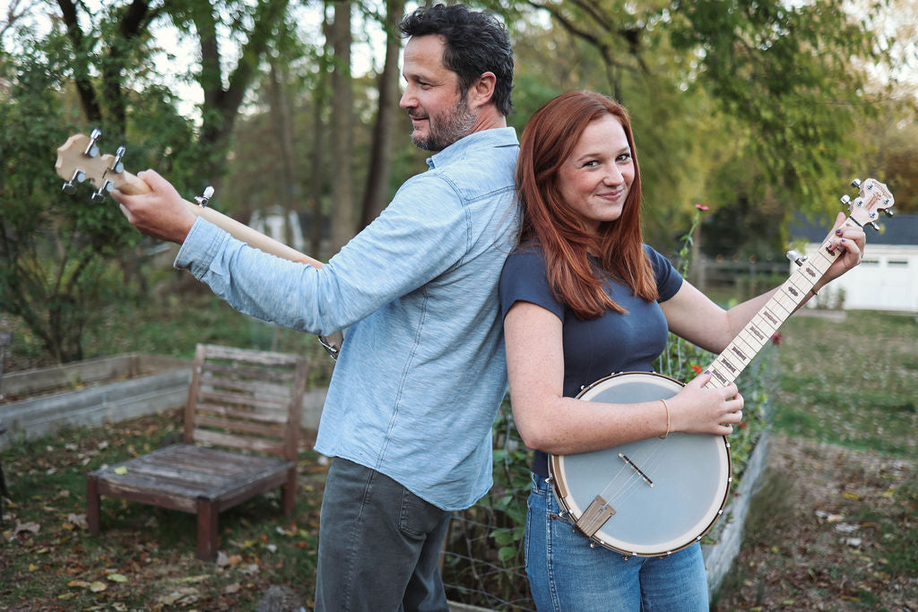 Man and woman playing banjos outdoors with trees and a bench in the background