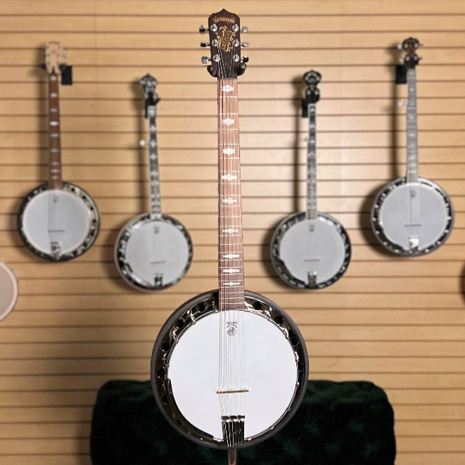Six string banjo with dark wood fingerboard and white head in front of a  Collection of banjos displayed against a wall with framed pictures.
