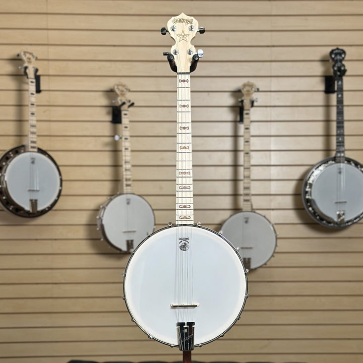 short scale 19 fret tenor banjo, natural blonde finish in front of wooden wall with banjo instruments hanging in the background. 