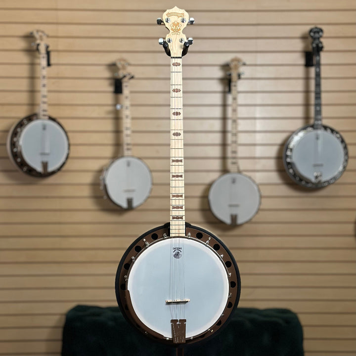 22 fret 4 string  banjo, natural blonde finish in front of wooden wall with banjo instruments hanging in the background. 