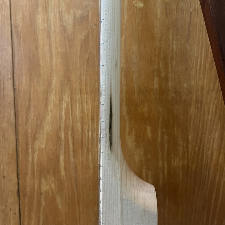 Close-up of a light-colored wooden banjo neck vertically against a warm brown wood-grain background.