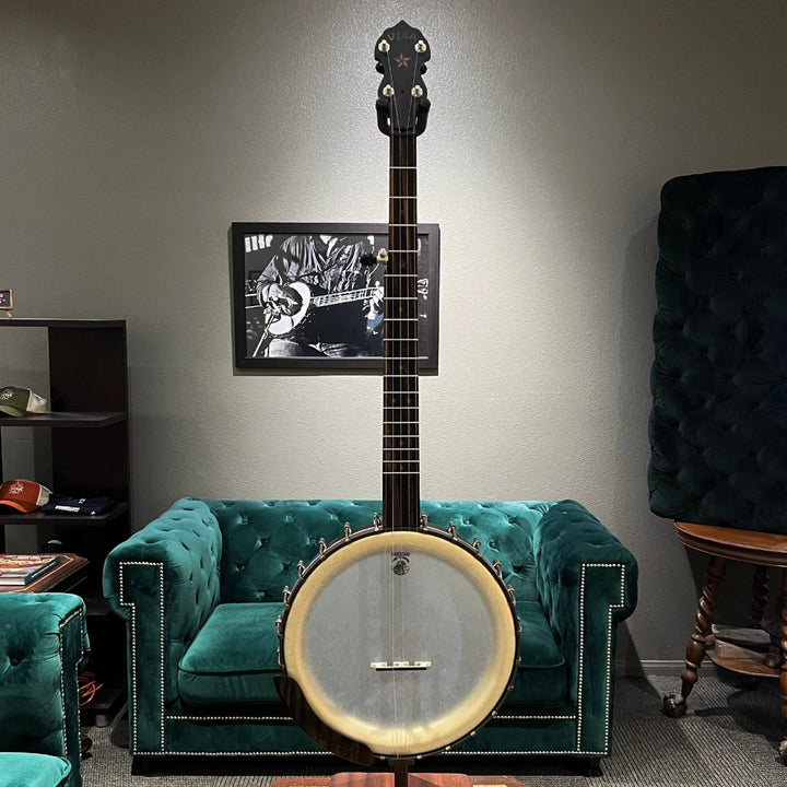 Banjo on a stand against a gray wall with a television displaying a person playing a guitar.