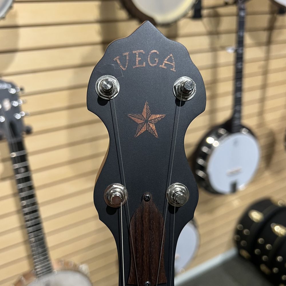 Close-up of a banjo peghead with Vega printed showing a visible crack in the black veneer at top left corner in front of wooden wall blurred banjos in background
