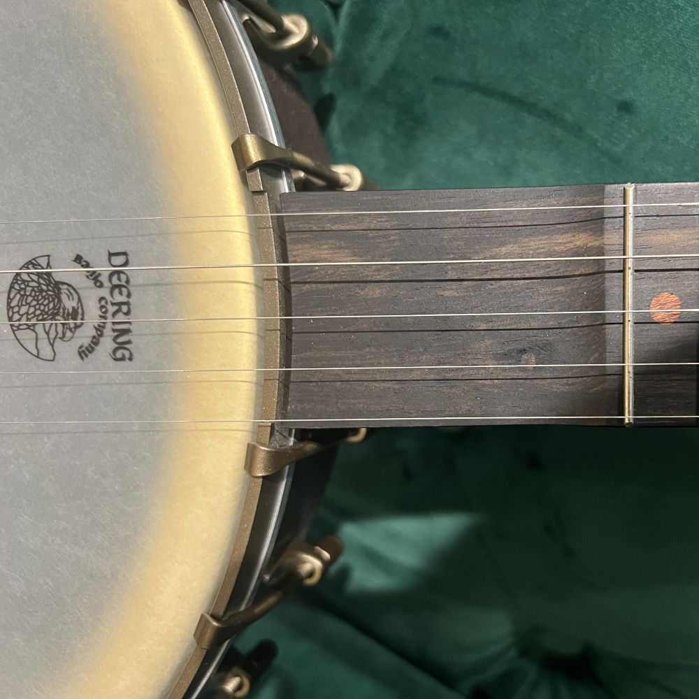 Close-up of a Deering banjo showing visible surface impressions and wood grain variations in the scooped fingerboard area, with the banjo head and Deering logo visible below. Green Velvet back drop.