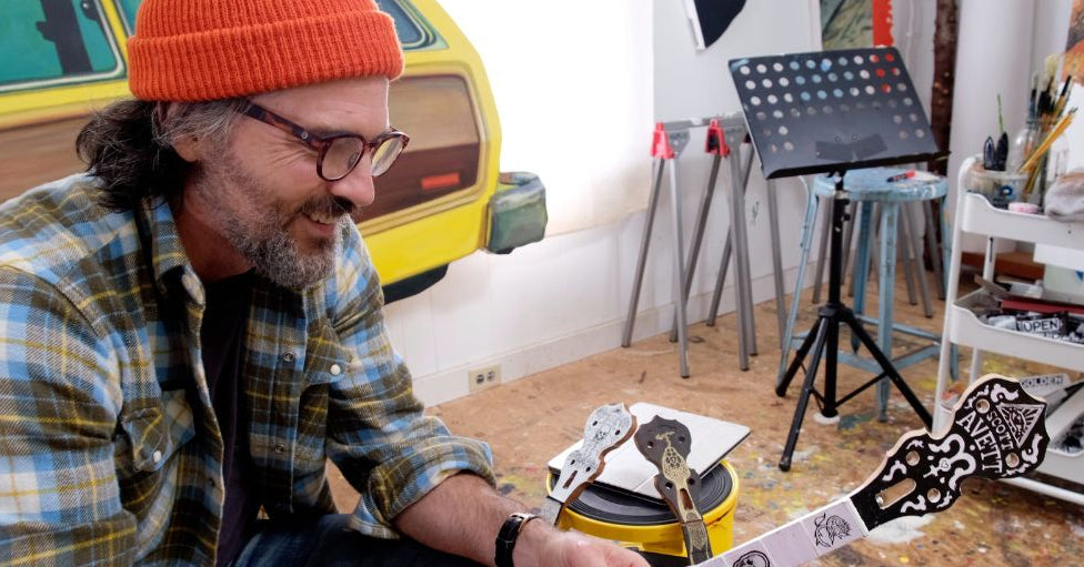 Scott Avett wearing an orange beanie and glasses sits in a creative studio, holding a white banjo neck. A yellow vintage car is visible in the background, along with music stands and an art cart.
